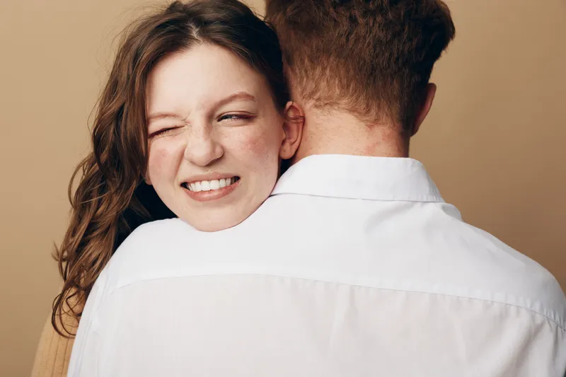 Happy young woman with curly hair smiles with closed eyes hugging man in white shirt. Intimate moment, love, relationship, affection, and emotional connection concept.