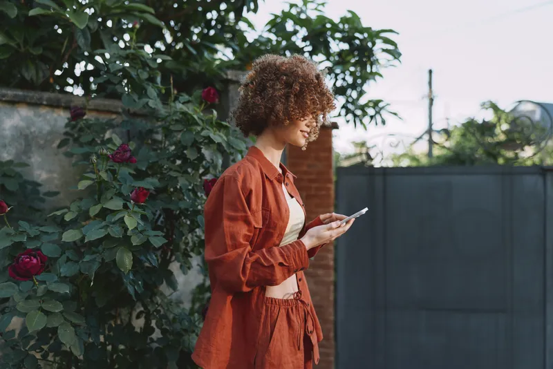 Young woman with curly hair checks her smartphone beside a rose hedge in a warm outdoor setting
