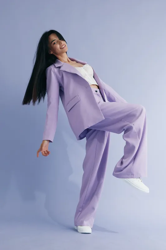 Young woman posing in lilac suit and white sneakers with long black hair against soft purple background, confident expression, modern fashion style, studio lighting.