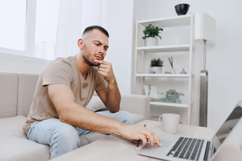 Thoughtful man working on a laptop at home, surrounded by plants and modern decor, reflecting creativity and focus. Perfect for themes of productivity and remote work.