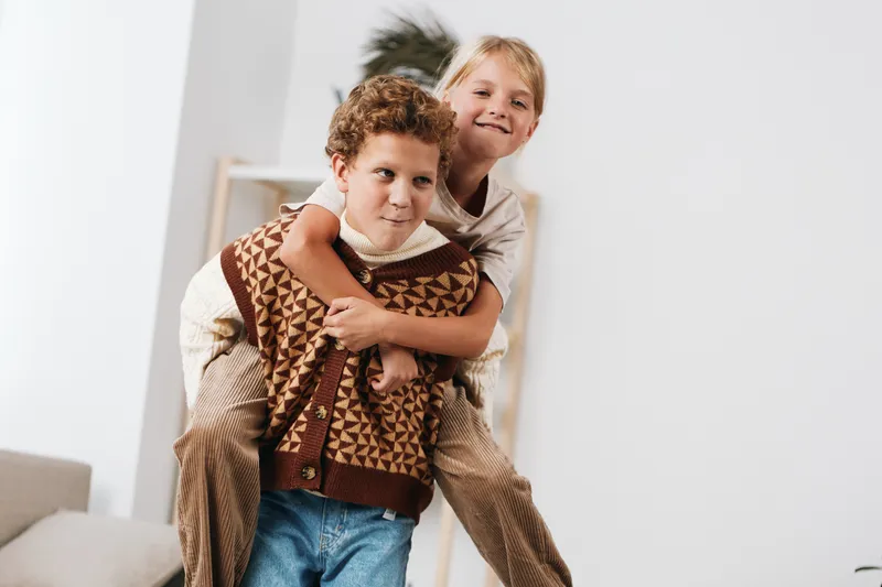 Two children share a playful piggyback moment in a bright home, smiling and close, wearing cozy knitwear and denim, with soft natural light creating a warm, candid scene of friendship and joy.