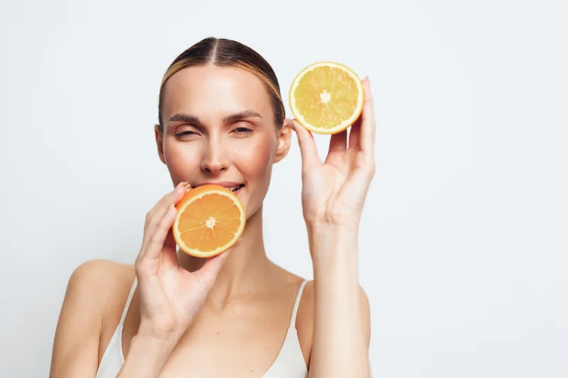 Woman skincare vitamin c citrus facial wellness self care in studio white background young woman holds orange halves by her face soft lighting for fresh hydration