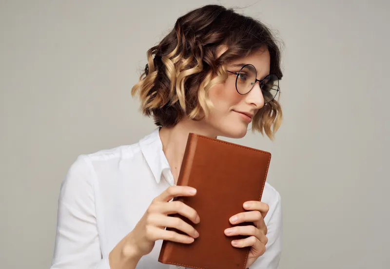 Женщина с вьющимися волосами в очках держит в руках книгу и работает в студии