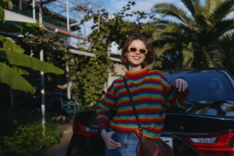 Young woman in striped sweater and sunglasses leaning on car trunk, smiling in tropical outdoor street. Casual outfit, crossbody bag, short hair, travel lifestyle mood.