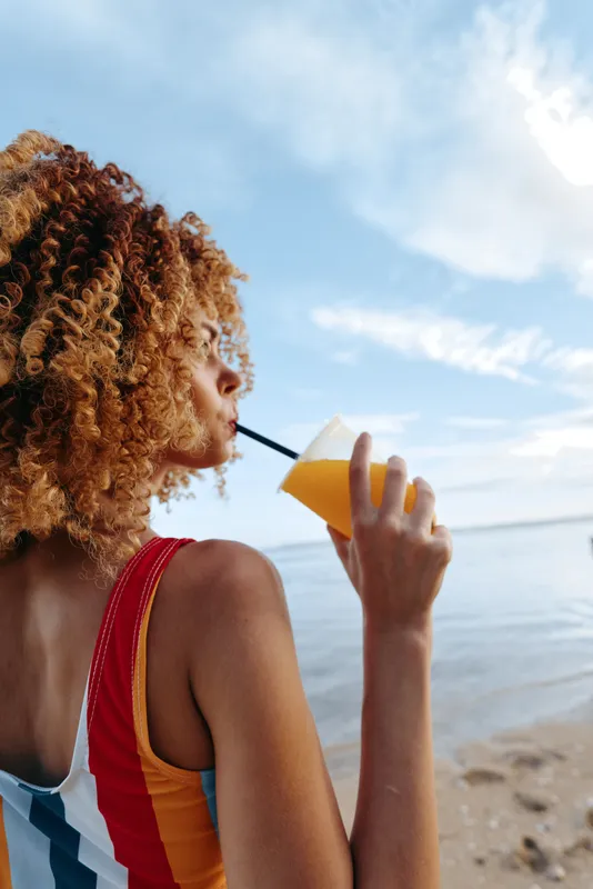 Young woman with curly hair drinks orange juice through a straw while standing on a sandy beach under a partly cloudy sky during a warm day.