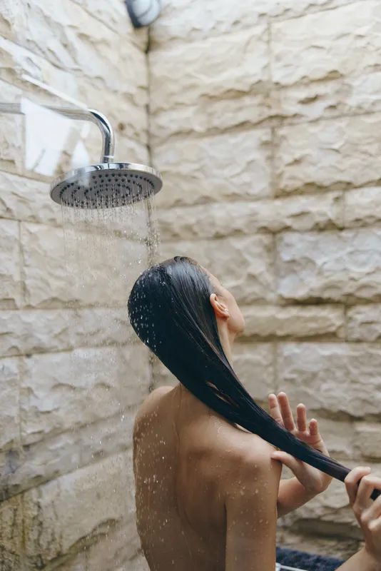 woman showering outdoors under rainfall showerhead, wet hair, natural stone wall, spa atmosphere, relaxation moment