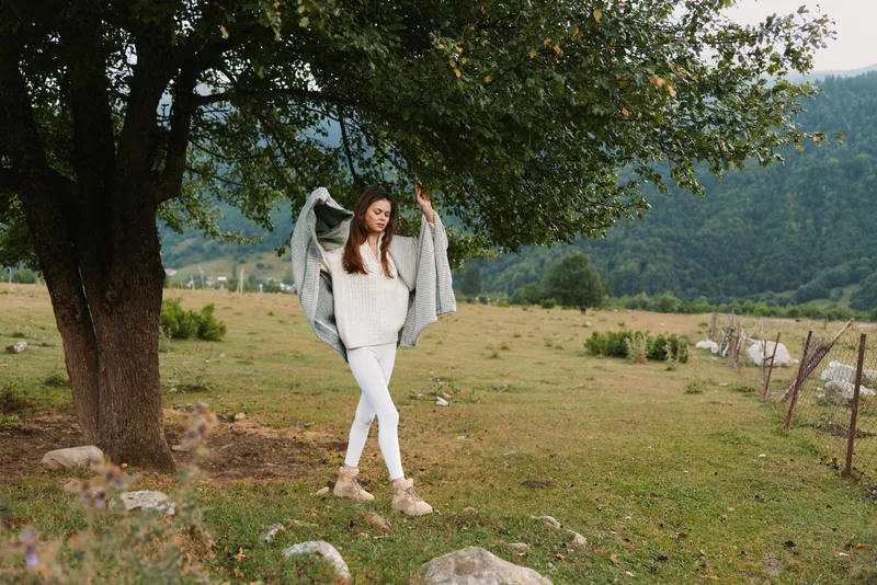 Young woman in white sweater and pants enjoys a relaxed outdoor moment beneath a large tree in a rural field, with a gentle breeze and peaceful countryside vibe.