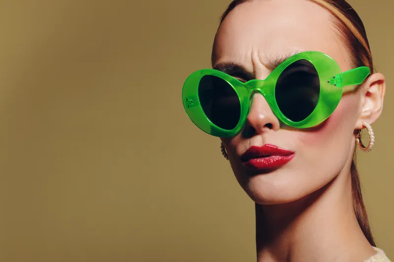 Close-up of a woman wearing large green sunglasses with red lipstick and hoop earrings. She looks serious against a plain beige background. Fashion and style concept.