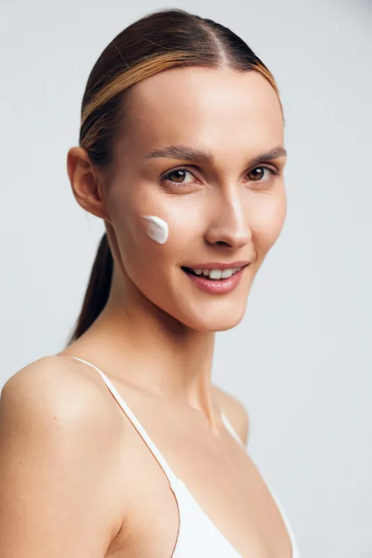 Young woman with smooth skin applies cream to her cheek in a bright studio, highlighting skincare routine, self care, and a calm, confident mood, with soft lighting that enhances texture and glow.