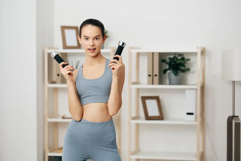 Fitness woman in a light blue sports set lifts dumbbells in a clean home gym, showing strength, balance and focus during an energetic, results-driven workout.