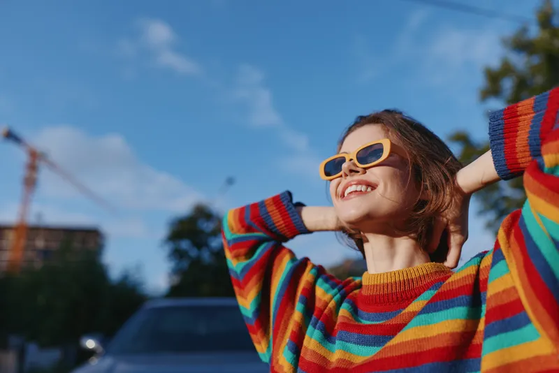Woman smiling in a striped sweater and yellow sunglasses outdoors in a city street, young model leaning back with hands behind head, sunlit casual outfit for lifestyle travel and fashion.