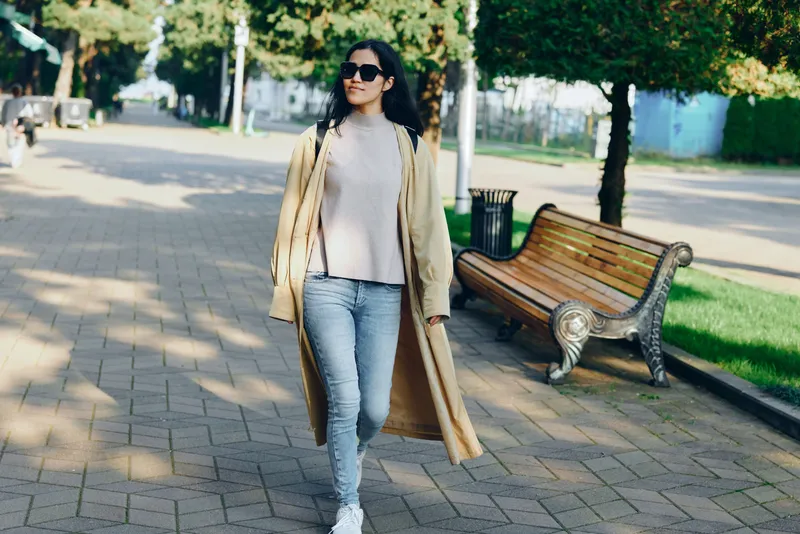 Woman in a long beige coat and jeans walks along a park path, wearing sunglasses, showcasing casual street fashion in a sunny outdoor setting with trees