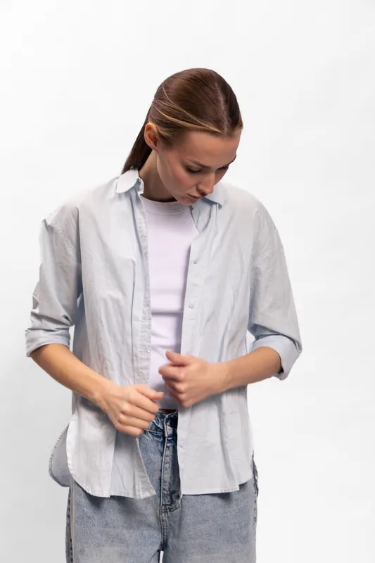 Young woman in casual outfit buttoning her light shirt while looking down, isolated on white background. Concept for fashion, style, and everyday clothing.