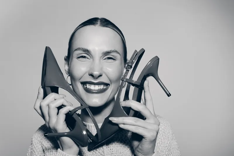 Smiling white woman with light skin holds high heel shoes near her face, showing joy and fashion style on plain background.