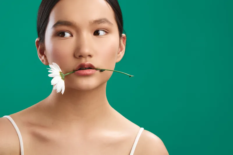 Asian woman with a flower between her lips presents a calm, artistic portrait against a vibrant green background, blending natural beauty with modern studio aesthetics.