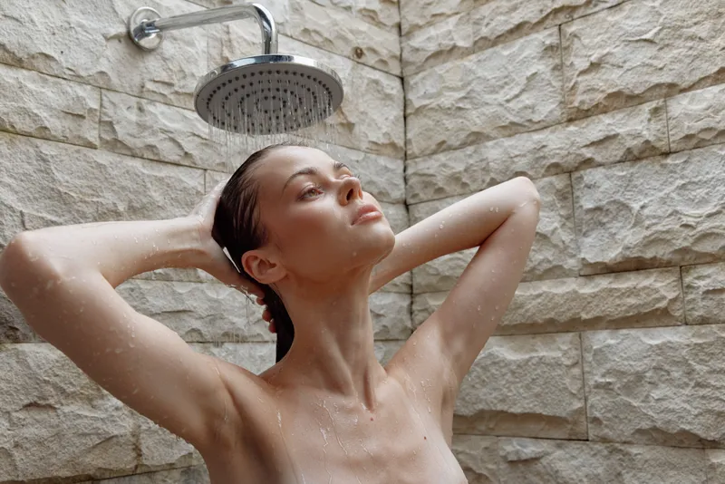 Young woman taking shower with hands behind head under rain showerhead against textured stone wall, enjoying relaxing moment in minimalistic bathroom interior.