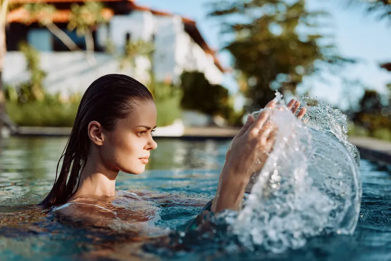 A carefree woman enjoys a sunny pool moment, water splashes as she playfully flicks. A bright, relaxed scene capturing leisure, outdoor lifestyle, and poolside calm.