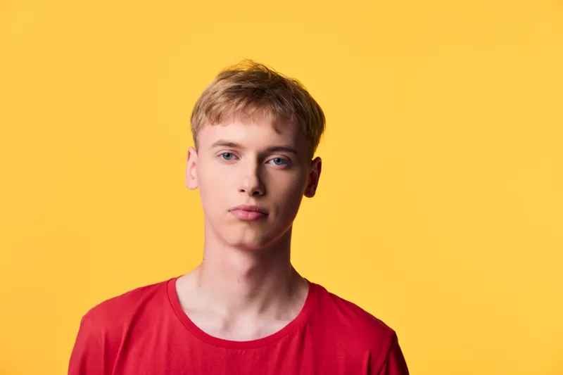 Young man in a red shirt stands against a bold yellow backdrop, showcasing contemporary casual style, simple composition with clean lighting, minimal distraction, suitable for fashion, lifestyle, or