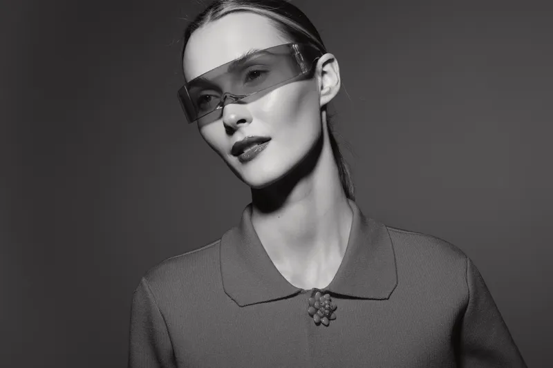 Stylish Caucasian woman with pale skin wears futuristic glasses and textured jacket in black and white studio portrait.