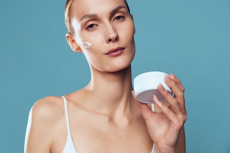 A young woman with clear skin holds a jar of face cream and applies a small amount on her cheek. She poses against a plain blue background in a white tank top. Concept for skincare, beauty, and