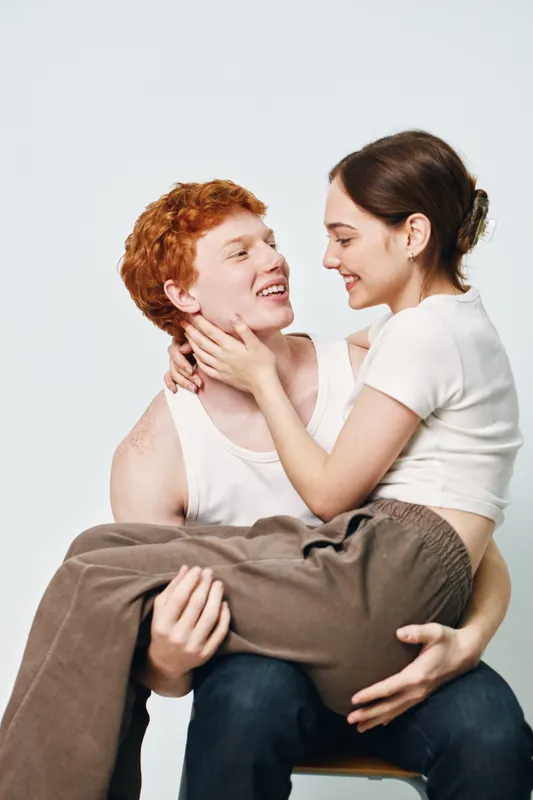 Couple in love smiling and embracing with woman sitting on man lap close to each other in casual clothes against white background. Young partners sharing happy moment together.