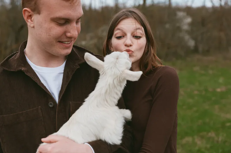 Casual outdoor scene capturing a couple with a friendly goat kid, sharing a tender moment in green fields, highlighting animal interaction, affection, and connection with nature