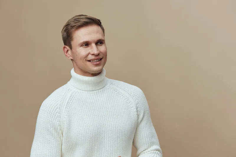 Smiling young man with light brown hair wearing cozy white knitted sweater looking away isolated on beige background, cheerful casual portrait. People lifestyle concept