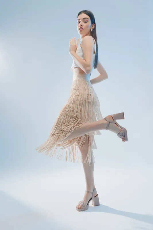 Young woman in a beige fringed skirt and white top poses elegantly on one leg in high heels. Studio shot on a light background. Concept of woman beauty fashion.