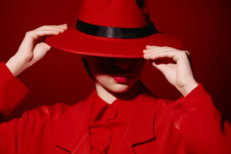 Stylish woman in vibrant red outfit partially covering face with wide-brimmed hat in matching color. Fashion photography emphasizing beauty, grace, and bold color contrast.