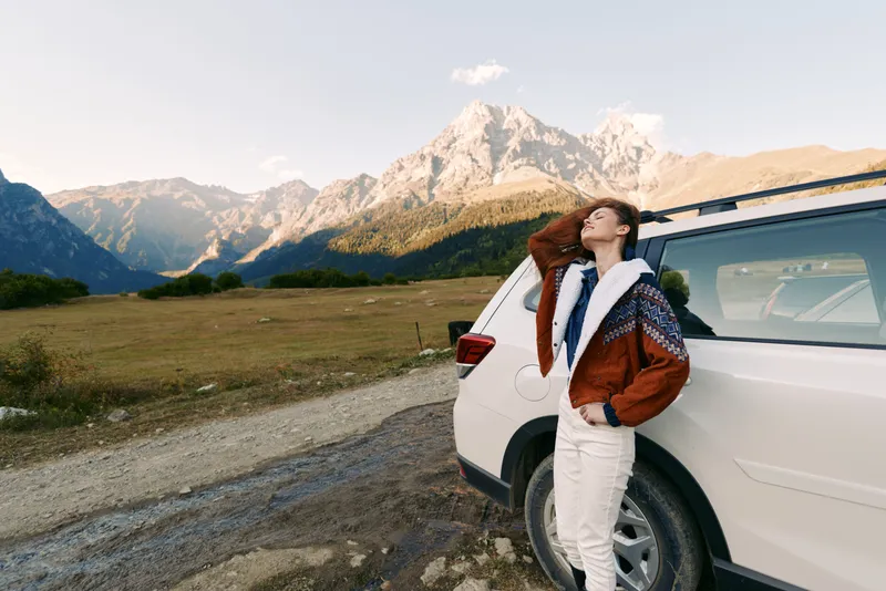 Woman by car at roadside with mountains in background, enjoying travel and nature during a scenic road trip, relaxed pose leaning on white SUV at sunset landscape.