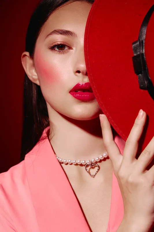Close-up portrait of a stylish woman with glossy red lips and pink blush holding a vibrant red hat. Concept of beauty creativity fashion, emphasizing glamor and high-end style.