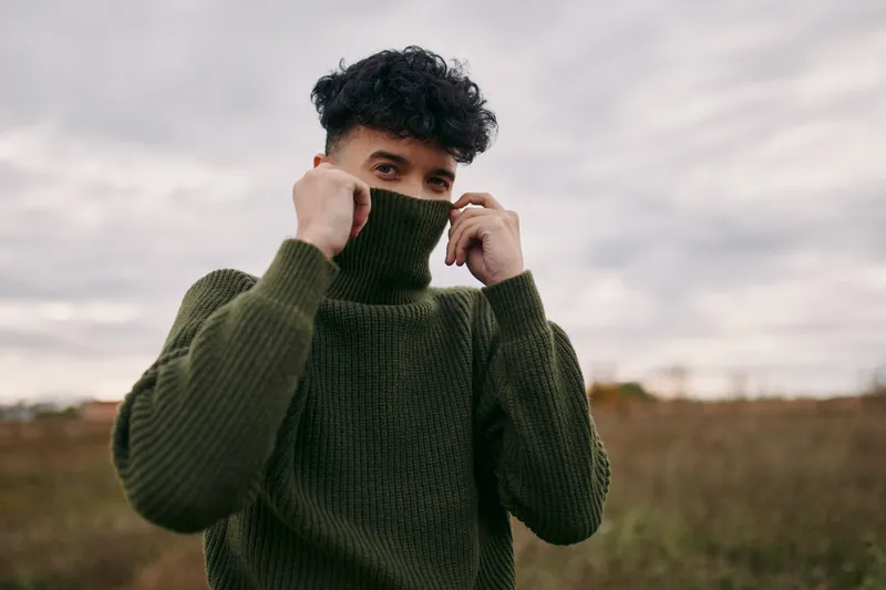 An authentic portrait of a young person outdoors, wearing a bulky green sweater, pulling up the collar for a candid reveal, conveying genuine mood and credible presence.