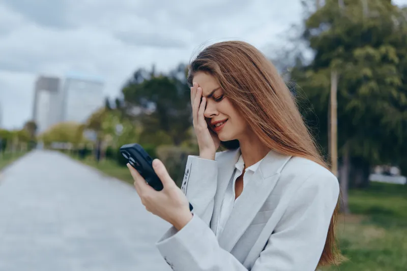 frustrated woman holding smartphone outdoors in gray blazer feeling stressed about finance and business crisis