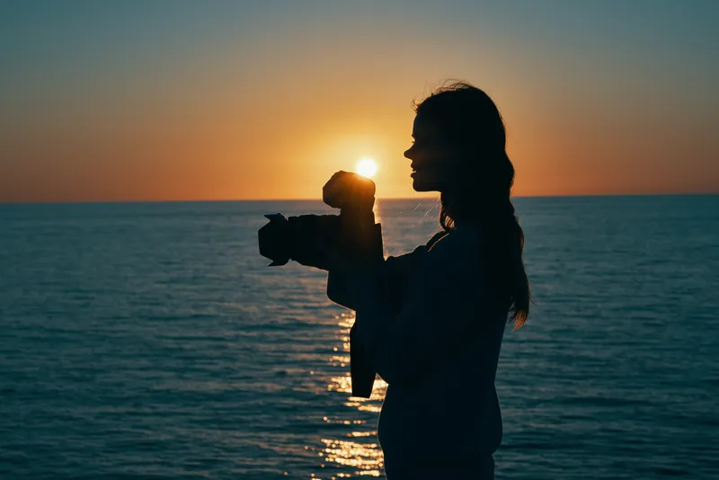 Женщина-фотограф с камерой на закате, на заднем плане море