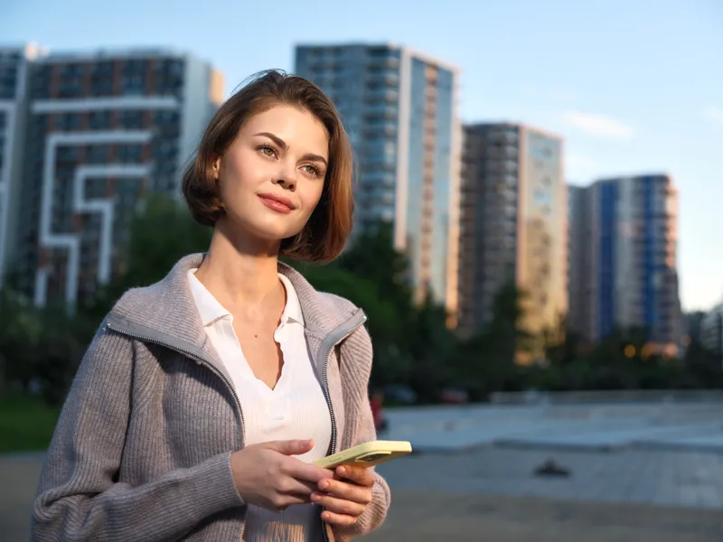 A confident woman stands outdoors in an urban park, holding a smartphone. She looks thoughtful and optimistic, suggesting focus and connectivity in a modern workday.