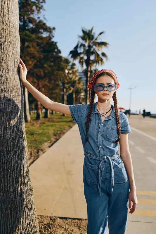 lifestyle woman in modern boho-western denim street style with sunglasses and braided hair, standing outdoors beside a tree under bright film color lighting