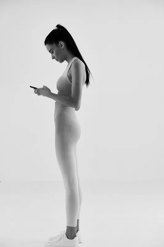Woman sport fitness standing in profile wearing light activewear focused on smartphone, isolated on white background in minimalistic studio setting.