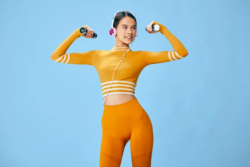 Young woman in orange activewear lifts small dumbbells while wearing pink headphones, smiling confidently against a clean light blue studio background, fitness concept.