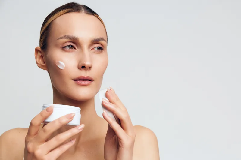 Young woman with clear skin applying white face cream on cheek. Beauty skincare concept with natural makeup and soft lighting on a plain white background. Self-care routine.