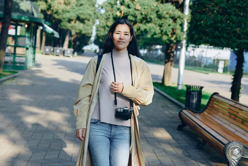 Casual dressed woman walks through a sunlit park, long cardigan flowing, small camera hung at neck, strolling along a paved path under leafy trees