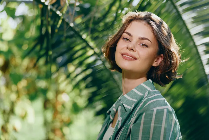 Young woman outdoors wearing a green striped shirt smiles softly, framed by lush tropical greenery and sunlit leaves.
