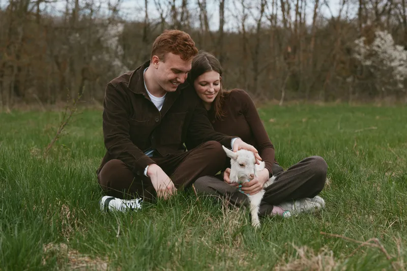 Couple sitting on a grassy field with a goat kid, enjoying outdoor life together. Warm light highlights affection, countryside simplicity, and wholesome lifestyle moment. Natural setting.