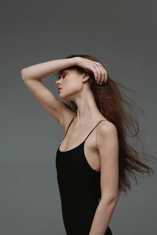 Elegant woman posing with flowing hair in a black dress, showcasing graceful movement and calm expression against a neutral gray background.
