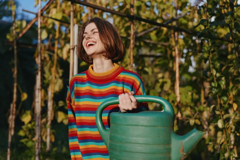 Woman with watering can in garden, smile bright while wearing a colorful striped sweater near greenhouse and vines. Young gardener enjoying sunlight, planting, potting and outdoor hobby activity.