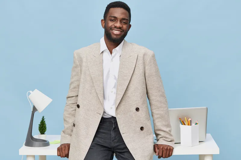 Confident smiling young African-American man in beige blazer and white shirt standing near office desk on pastel blue background, with laptop, stationery, and lamp, professional business casual look. 