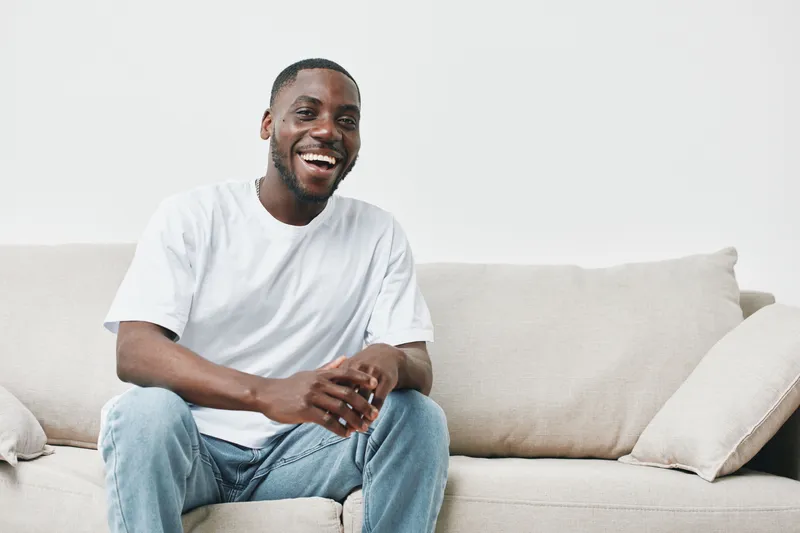 Cheerful young Black man in casual white t shirt sitting on a beige couch, expressing joy and positivity in a modern, minimalist living room