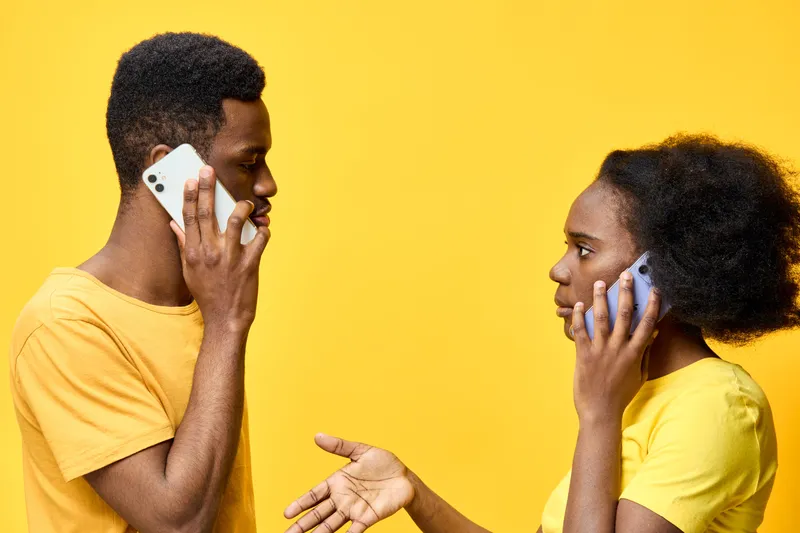 A young African American couple communicating with smartphones on a bright yellow background, expressing emotions through body language and facial expressions