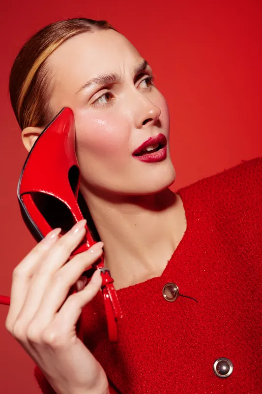 Young woman in red outfit holds red high heel shoe near face against red background. Fashion concept with bold colors, style, and confident female model in studio lighting.