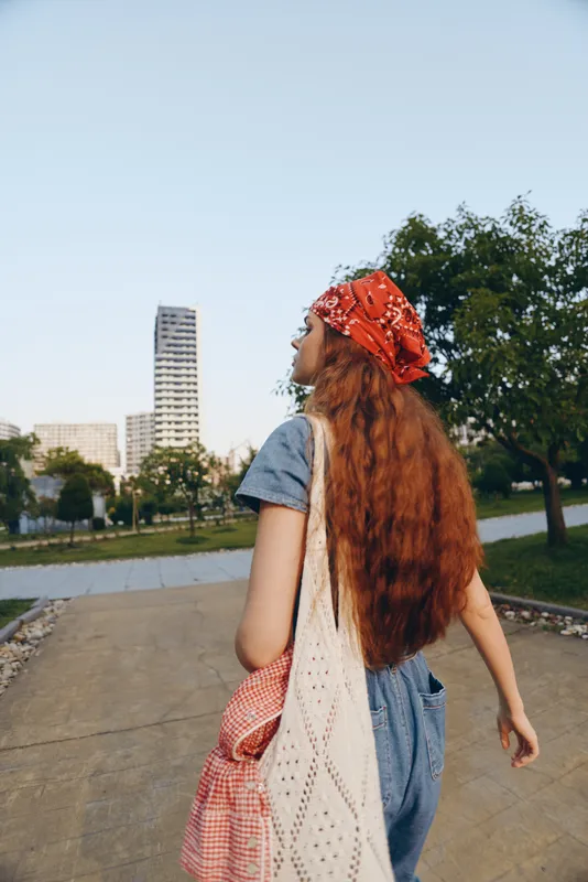 young woman with long red hair wearing a red headscarf and casual blue dress walking outdoors in a park under clear sky
