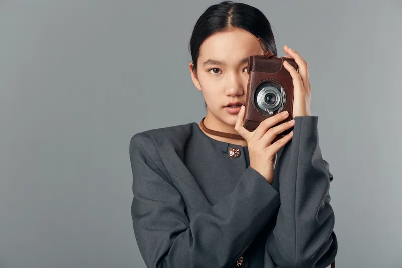 Young woman holding vintage camera close to face posing for portrait on grey background. Female model with photographic equipment wearing dark jacket captures picture in studio.